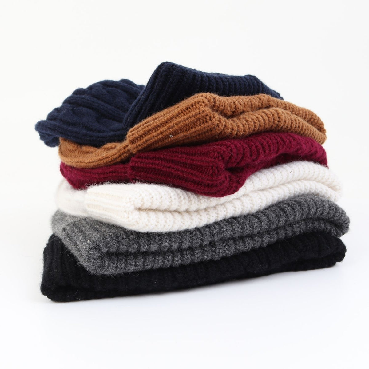 A stack of five folded cable knit beanies in navy, tan, burgundy, cream, and charcoal gray, photographed on a clean white background.