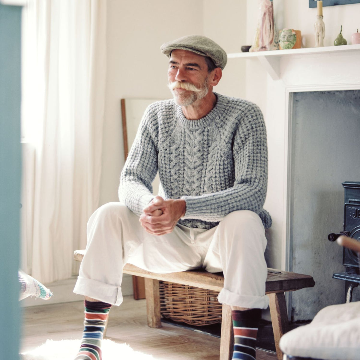 An older man with a white mustache sits on a wooden bench in a cozy room, wearing a chunky cable-knit light gray sweater, white trousers, and colorful striped socks.