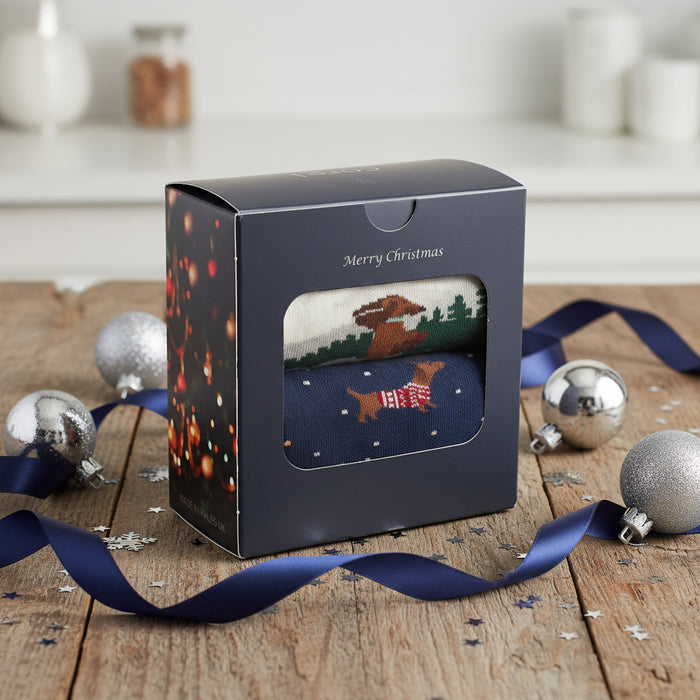 A gift box with a Christmas theme featuring a dachshund design, placed on a wooden surface with holiday decorations in the background.
