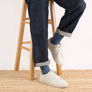 A person seated on a wooden stool wearing dark blue jeans with rolled cuffs, blue socks, and white low-top sneakers, photographed against a plain light background.