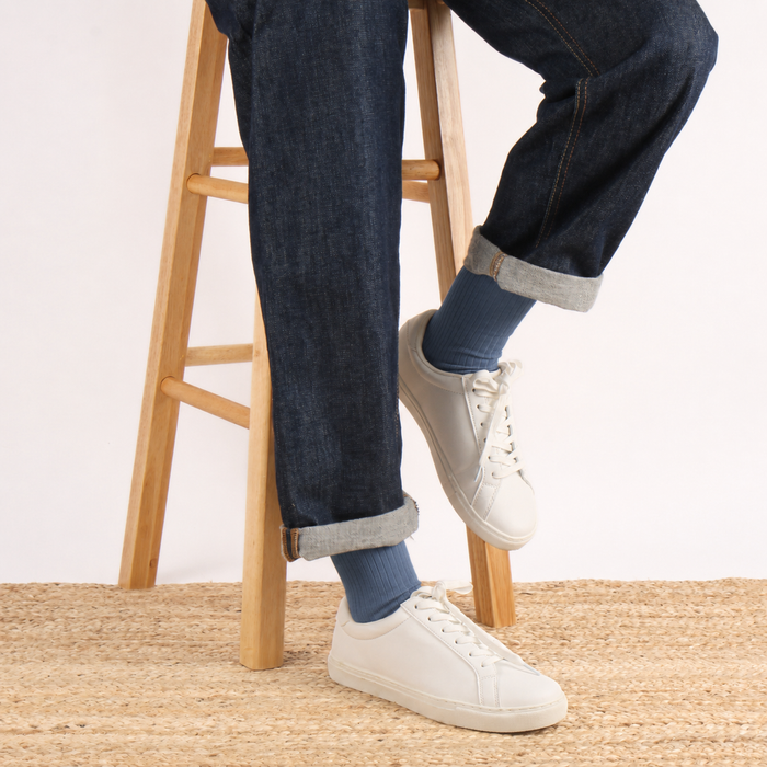 A person seated on a wooden stool wearing dark blue jeans with rolled cuffs, blue socks, and white low-top sneakers, photographed against a plain light background.