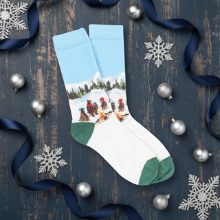A pair of kids' socks with a winter campfire scene featuring bears in blue and green colors, presented on a wooden surface with Christmas decorations around.