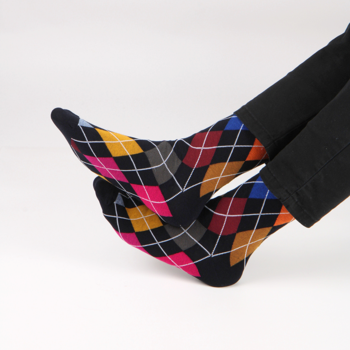 A pair of black socks with a colorful argyle pattern in pink, yellow, blue, red, and gray, worn on feet with black pants against a plain white background.