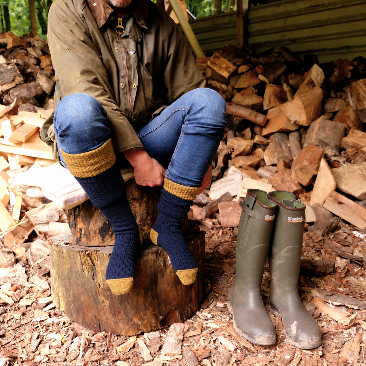 A person sits on a tree stump outdoors wearing dark blue knit socks with mustard yellow cuffs and toes, blue jeans, and a brown jacket. A pair of green rubber boots sits on the ground beside them, with stacked firewood and a rustic outdoor setting in the background.