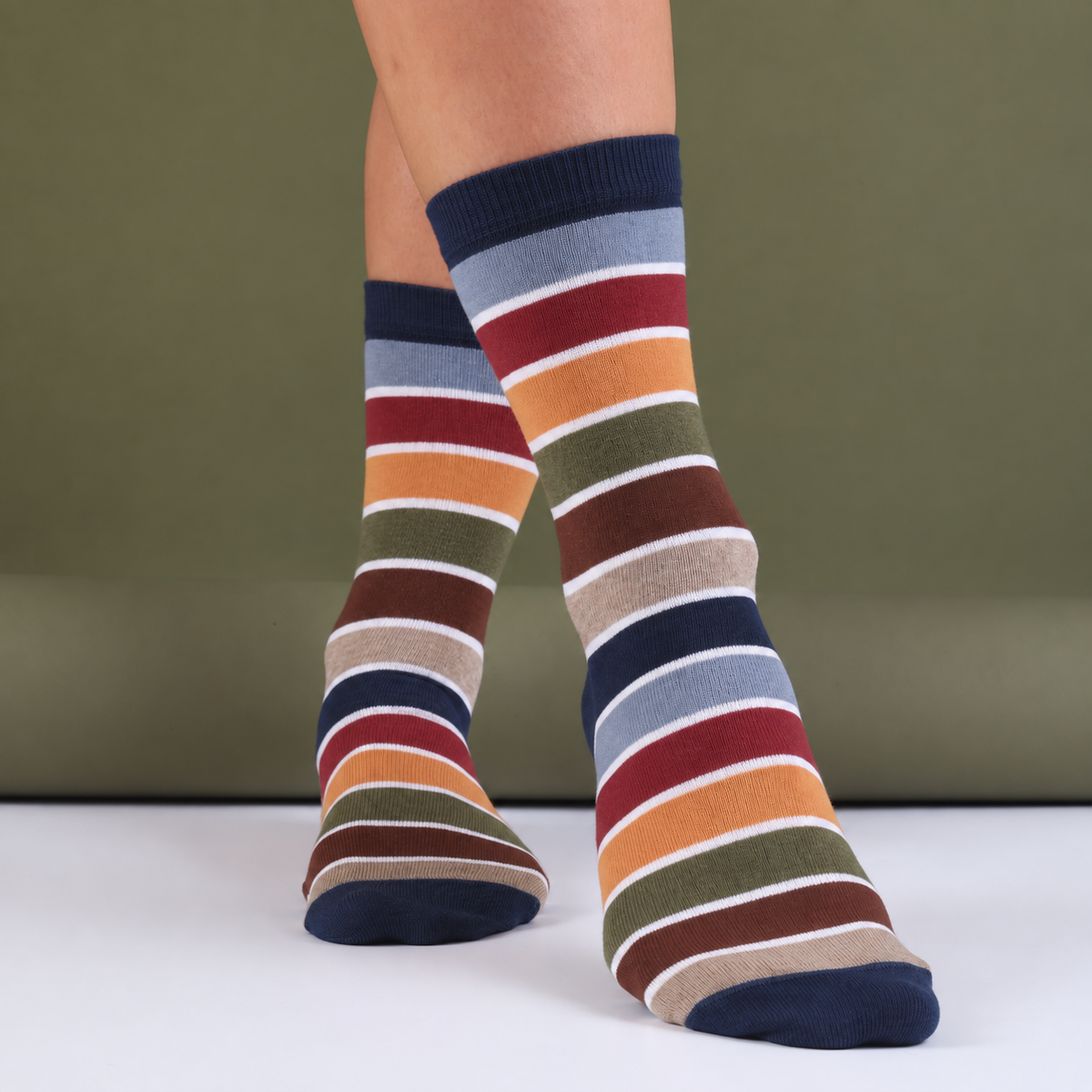 A pair of crew socks with horizontal stripes in navy, light blue, red, orange, olive green, brown, beige, and white, worn on a person's legs against a muted green background.