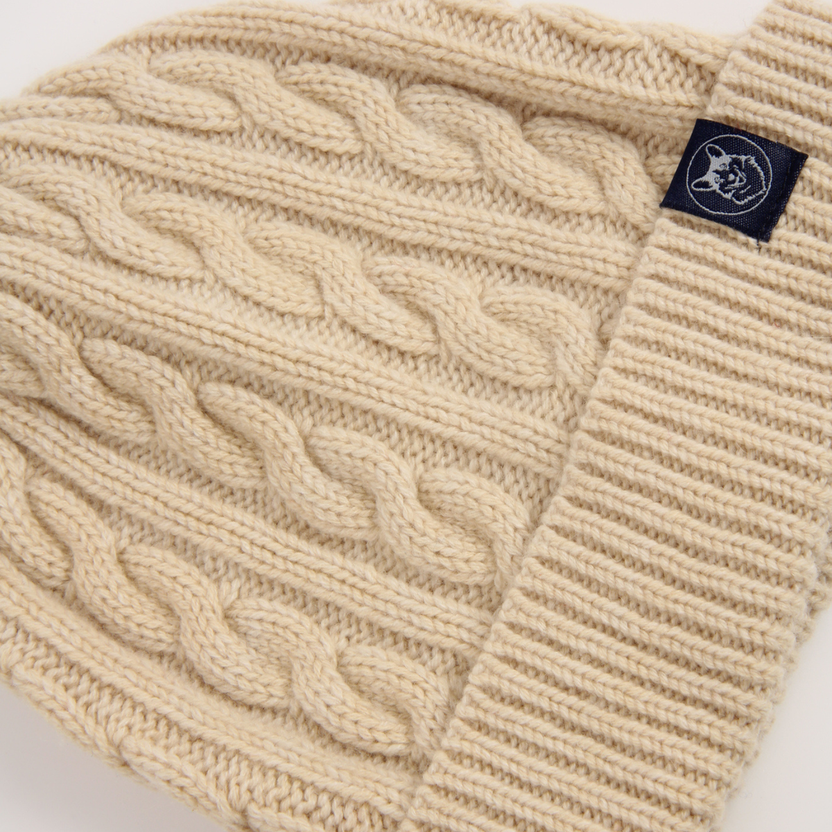 Close-up of a cream-colored cable-knit beanie with a ribbed cuff and a small navy blue brand tag on the side.
