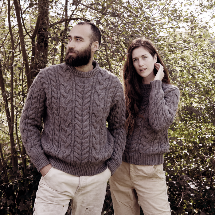 Two people wearing matching cable knit sweaters standing in a forest.