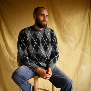 Man wearing a gray argyle sweater sitting on a wooden stool against a beige backdrop
