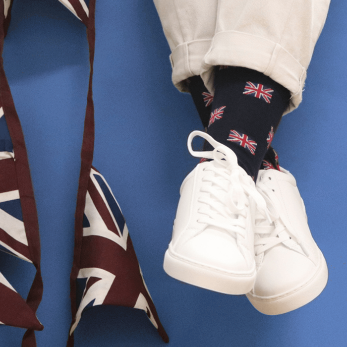 A pair of navy blue cotton socks with the Union Jack pattern, displayed on a pair of white sneakers against a blue background with a Union Jack tie in the corner.