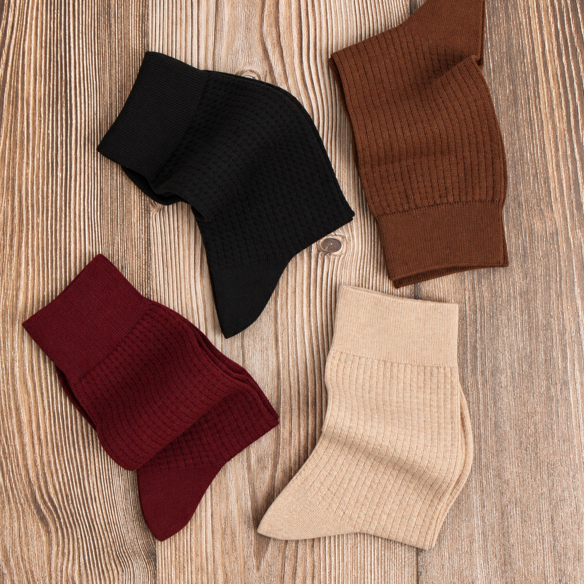 Four textured ankle socks in black, brown, burgundy, and beige, arranged on a light wood grain background.