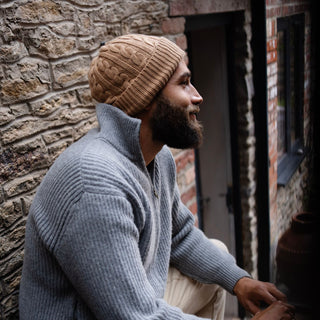 man wearing a cable cashmere beanie in colour brown sugar sitting outside on a step.
