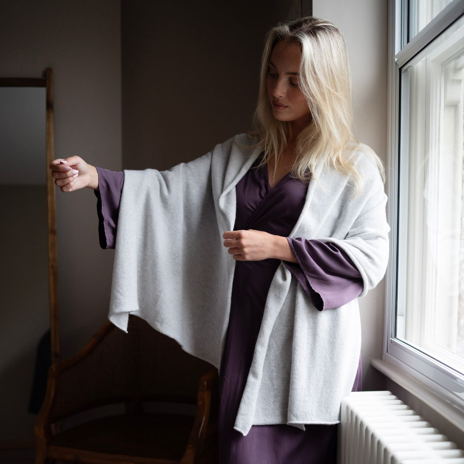 A woman stands by a window wearing a light gray shawl draped over a dark purple satin slip dress. She is in a dimly lit room with a wooden chair and mirror visible.
