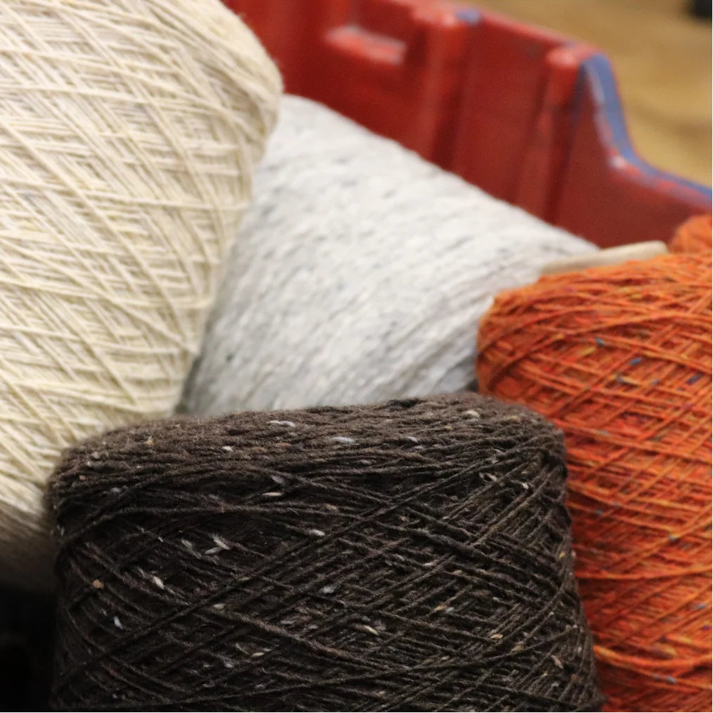 Close-up of several skeins of yarn in different colors, including cream, light gray, rust orange, and dark brown, with a red plastic container visible in the background.