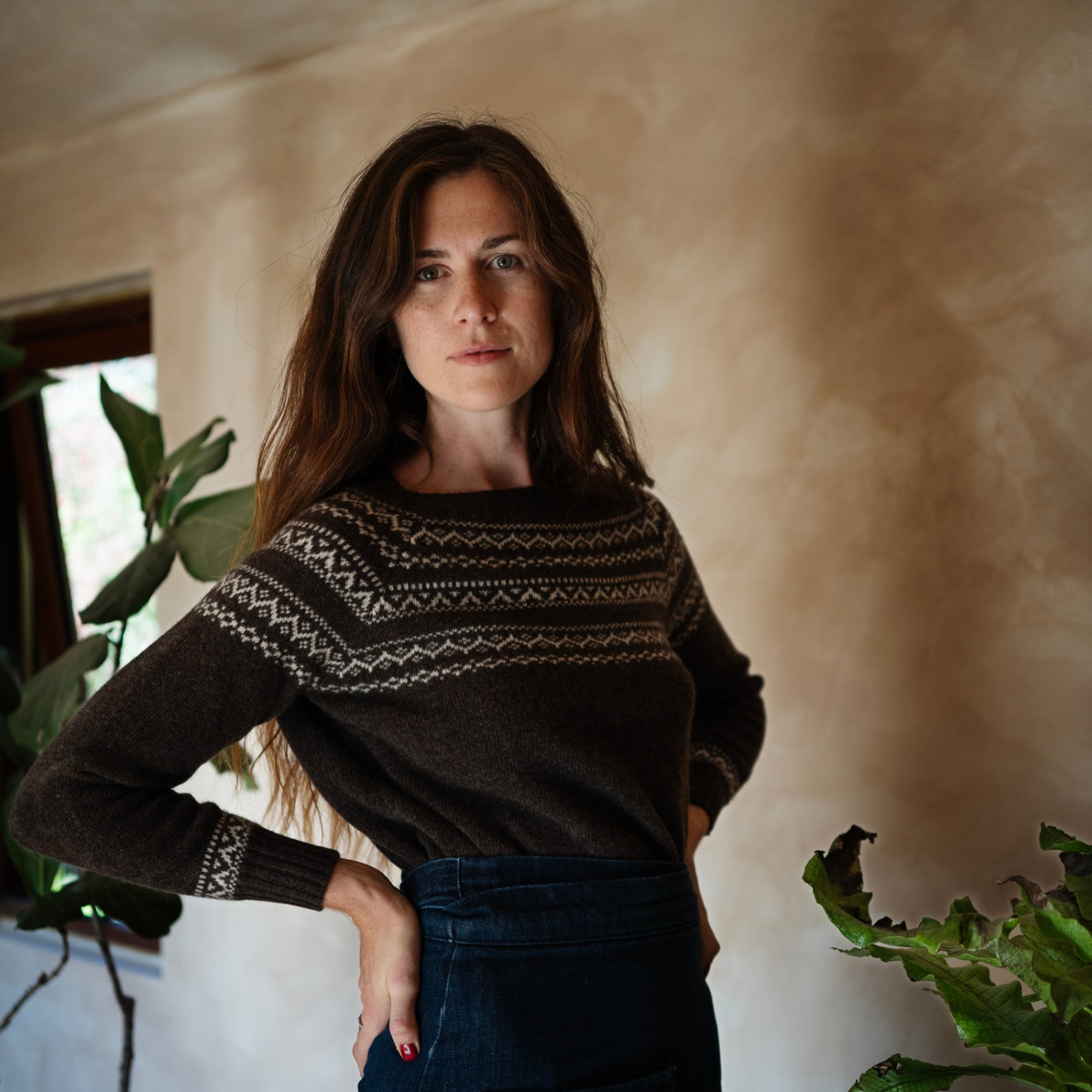 A woman wearing a dark brown knit sweater with a cream geometric pattern across the chest and cuffs, paired with dark blue jeans, standing in a warmly lit room with a textured beige wall and indoor plants.