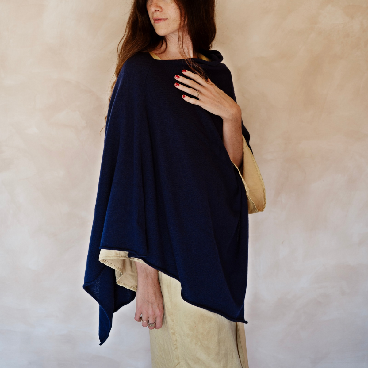 A woman wearing a navy blue wrap poncho with a beige lining, styled over a cream-colored dress against a textured beige background.