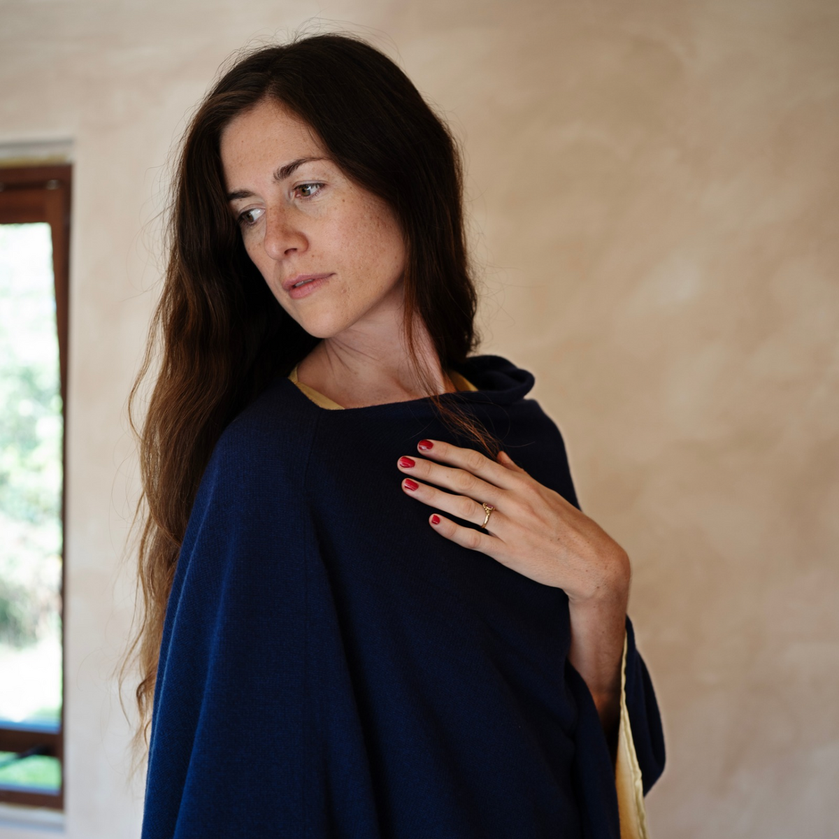A woman with long dark brown hair wearing a navy blue hooded poncho, standing against a textured beige plaster wall.