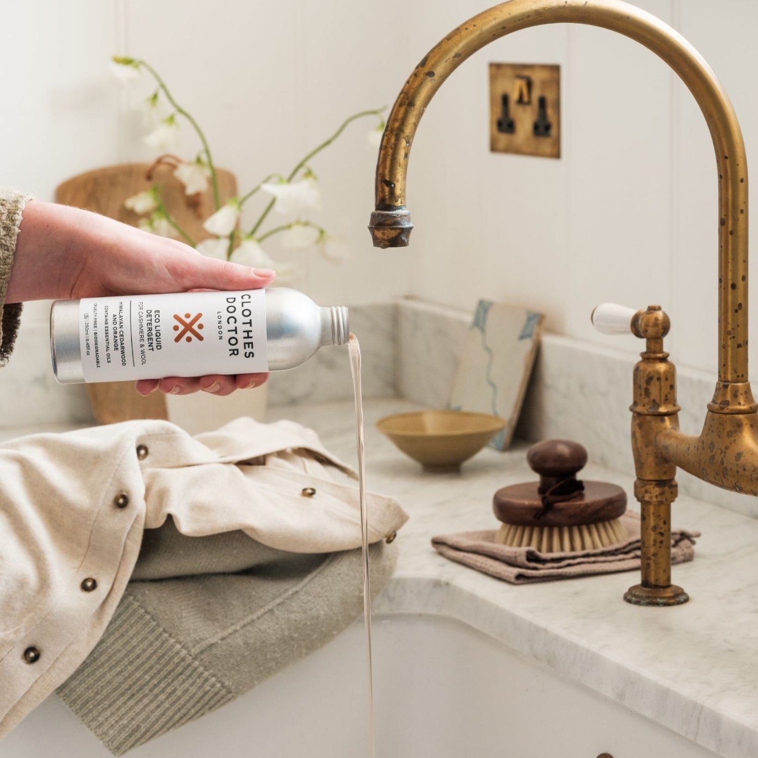 A bottle of Eco Wash for Cashmere & Wool detergent being held over a sink, with a garment laid out on the sink's edge, and a brush and soap dish beside the sink.