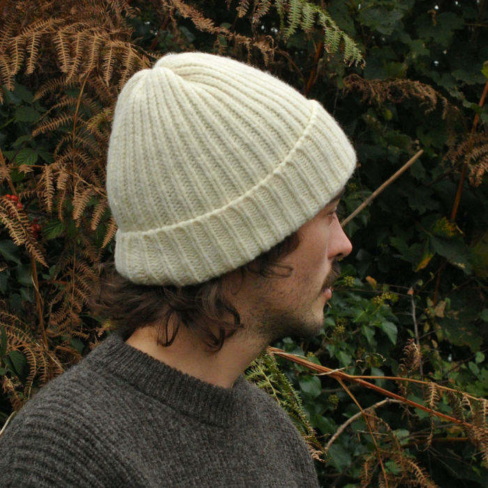 Person wearing a ecru knit beanie and sweater in front of green foliage