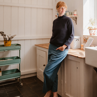 Person standing in a kitchen with white cabinets and a wooden countertop wearing a navy British wool roll edge sweater