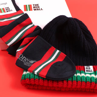 A set of Red Wall branded merchandise including a navy beanie with red and green stripes and a pair of matching socks, presented on a red background.