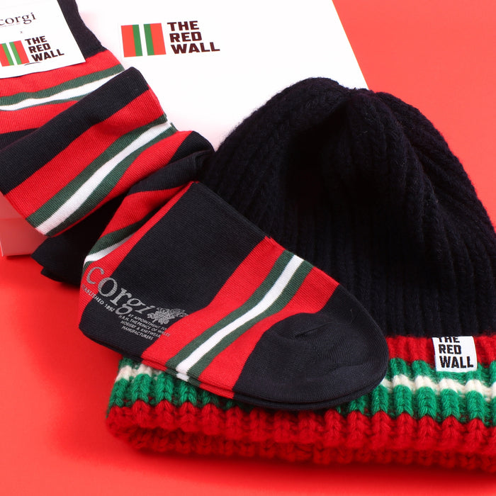 A set of Red Wall branded merchandise including a navy beanie with red and green stripes and a pair of matching socks, presented on a red background.