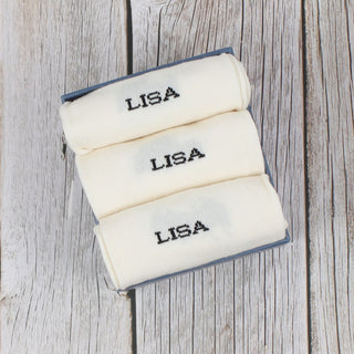 Three folded personalized socks with the name 'LISA' knitted on the side, displayed on a wooden surface.