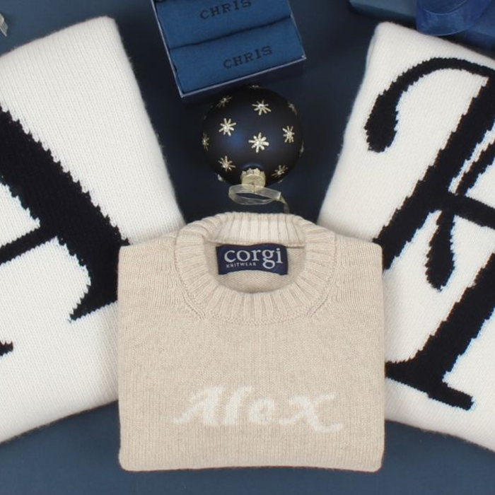 Beige sweater with 'Alex' knitted on it, placed on a dark surface with decorative items.