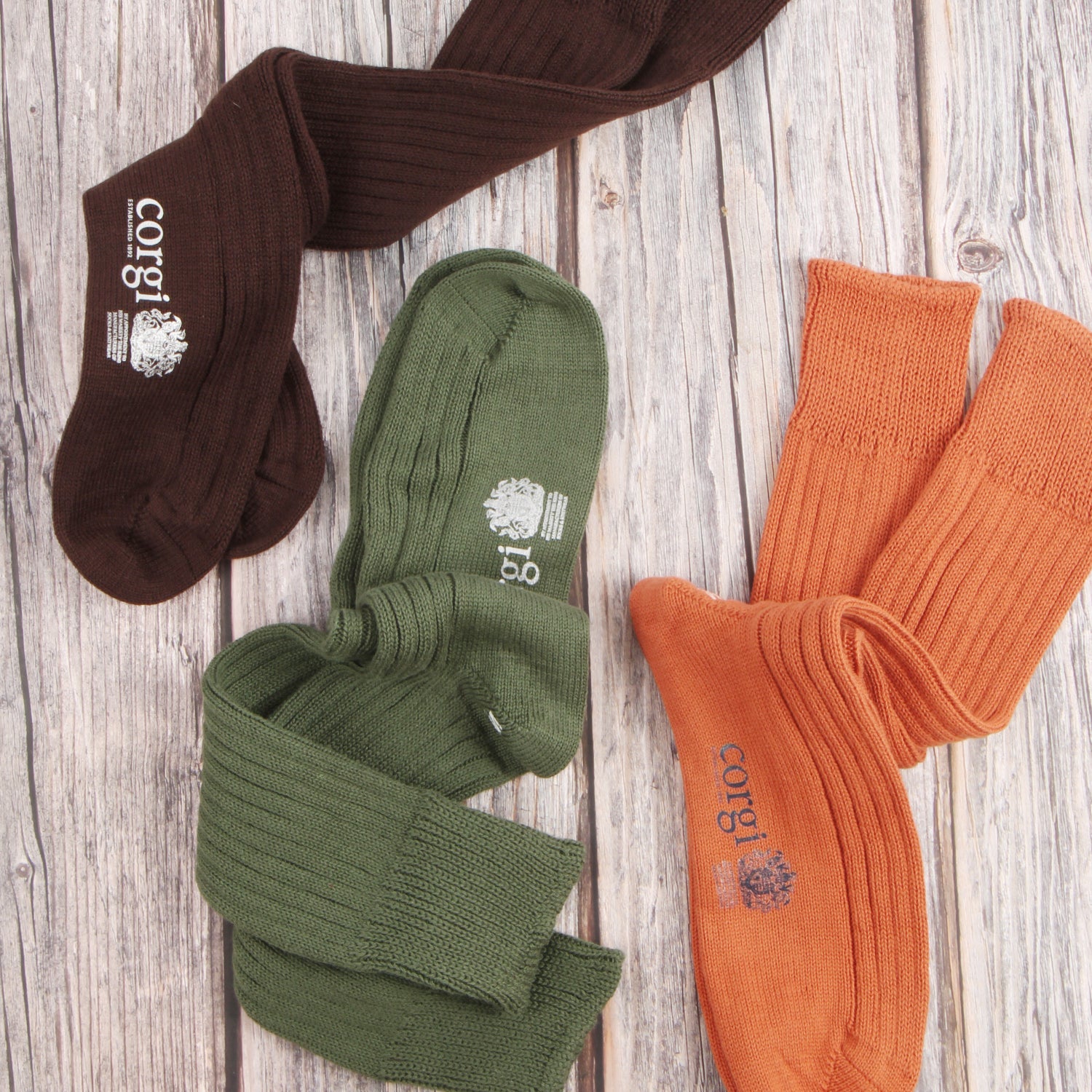 Three pairs of ribbed crew socks in brown, olive green, and orange, arranged on a light wood plank background. Each sock features a white printed logo with the text 'CORGI' and a crest.