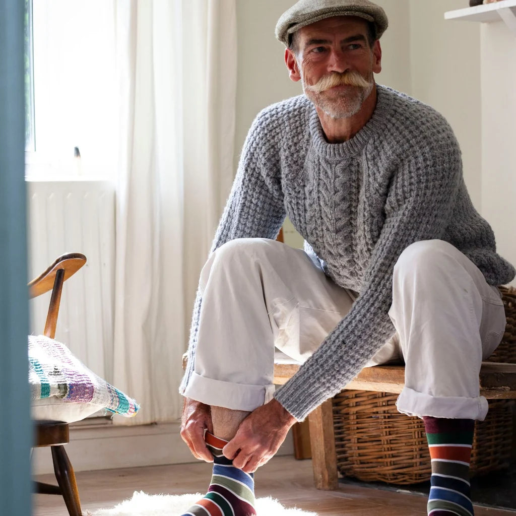 An older man with a white mustache sits on a wooden bench in a bright room, wearing a chunky gray cable-knit sweater, white trousers, and colorful striped socks while adjusting one sock.