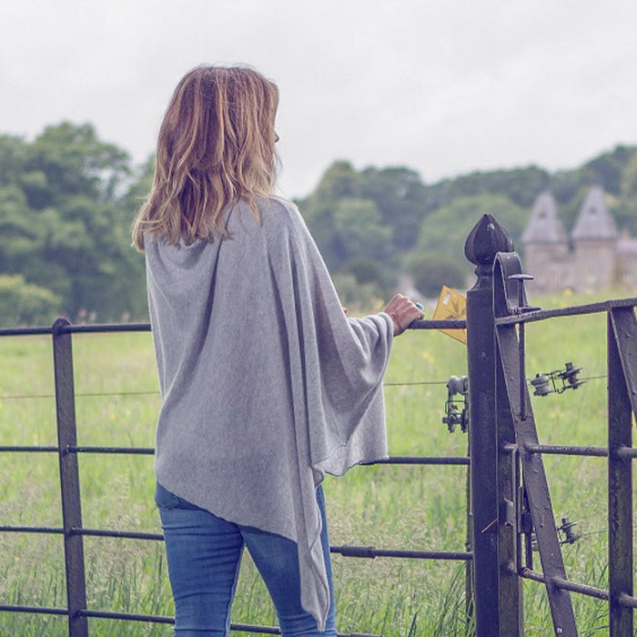 Woman in a gray poncho standing by a gate in a field with trees and buildings in the background