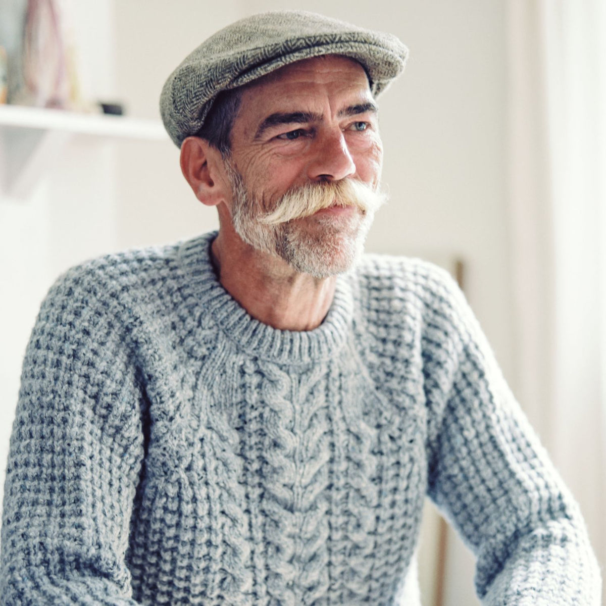 An older gentleman with a white beard and mustache wearing a gray cable knit wool sweater and a matching flat cap.