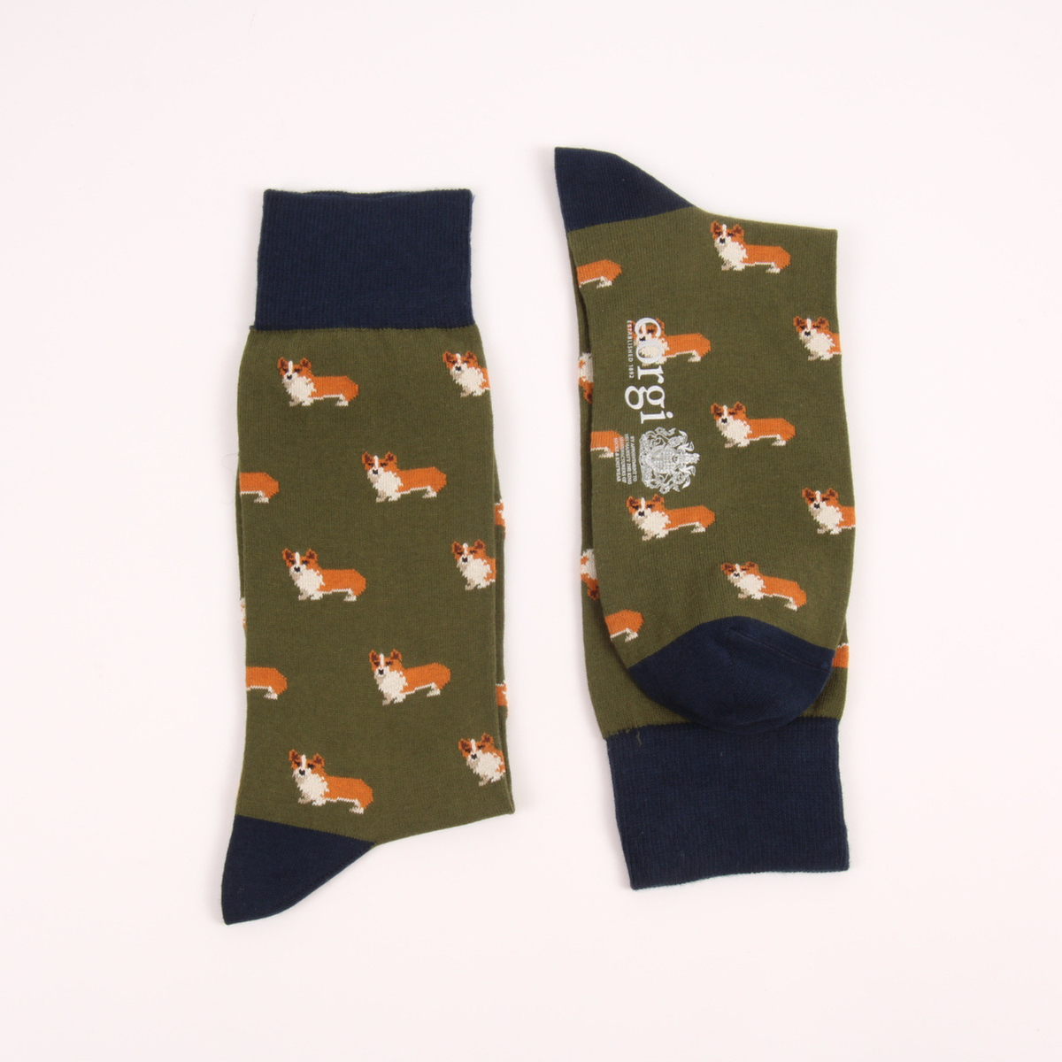A pair of crew socks laid flat on a light off-white background. The socks feature an olive green body with a repeating pattern of small Pembroke Welsh Corgi dogs in orange, white, and black, with solid navy blue cuffs, heels, and toes.