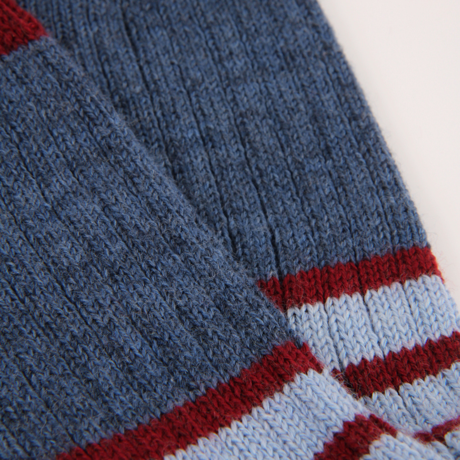 Close-up of a ribbed knit sock in medium blue with horizontal stripes of red and light blue near the cuff, shown against a plain light background.