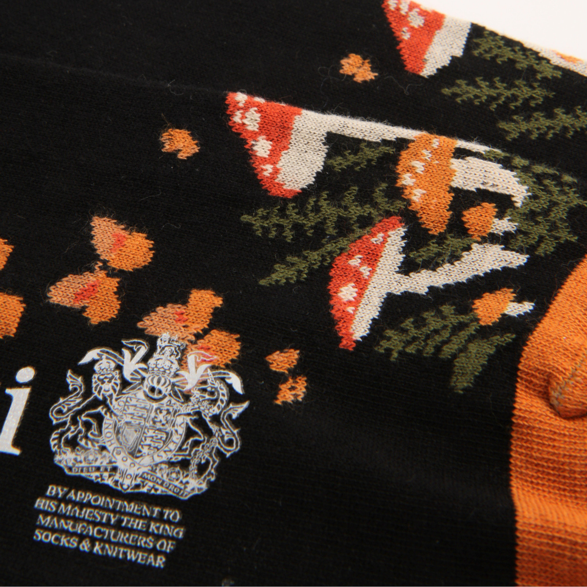 Close-up of a black knit sock featuring a colorful printed pattern of red-and-white mushrooms, green foliage, orange leaves, and a silver crest with text reading 'By Appointment to His Majesty the King Manufacturers of Socks & Knitwear.'.