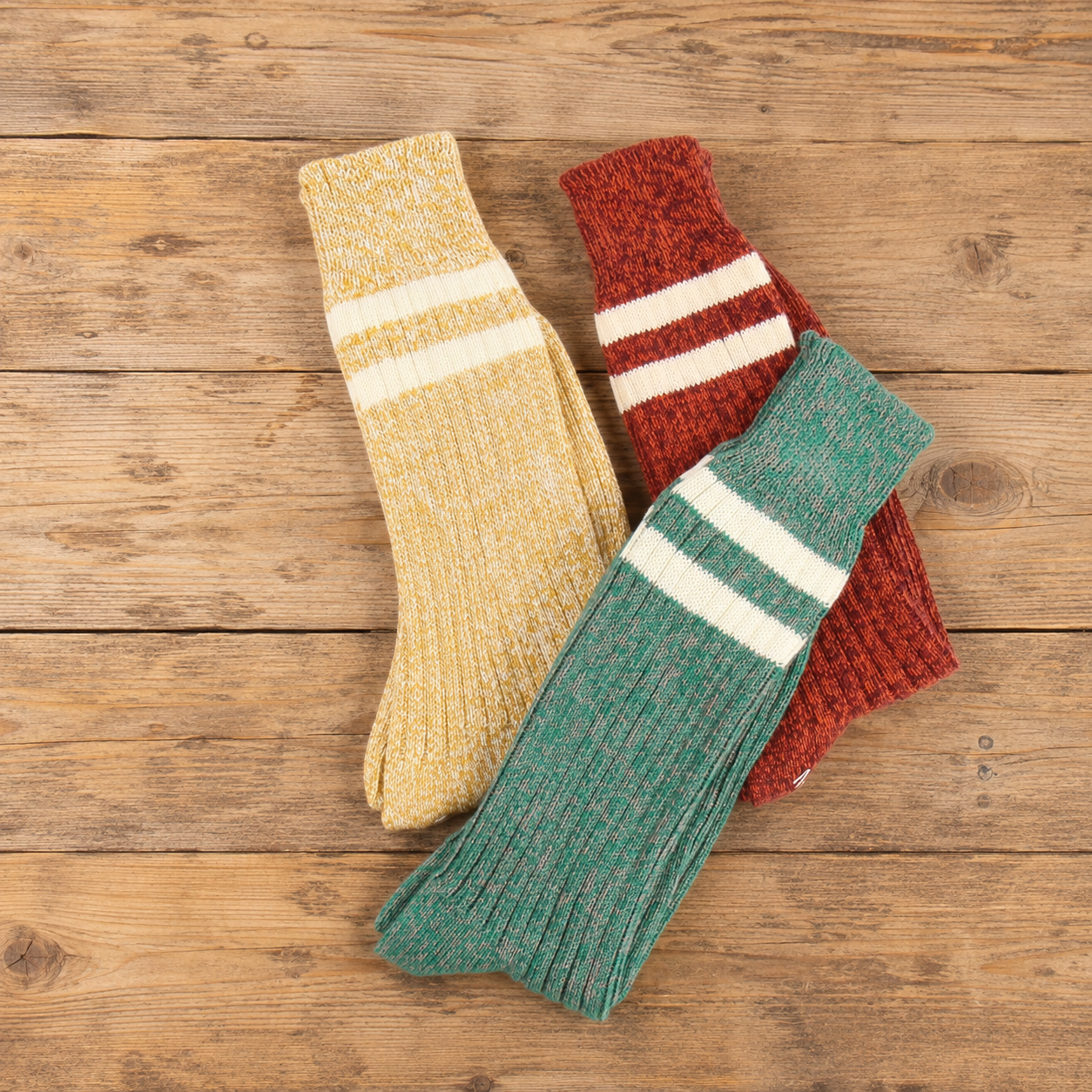 Three pairs of ribbed knit crew socks in beige, red, and green, each with two white horizontal stripes near the cuff, arranged on a rustic wooden surface.