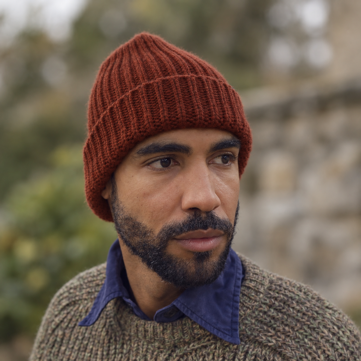 A close-up portrait of a man wearing a rust-brown ribbed knit beanie and a textured knit sweater over a blue collared shirt, with a softly blurred outdoor background.
