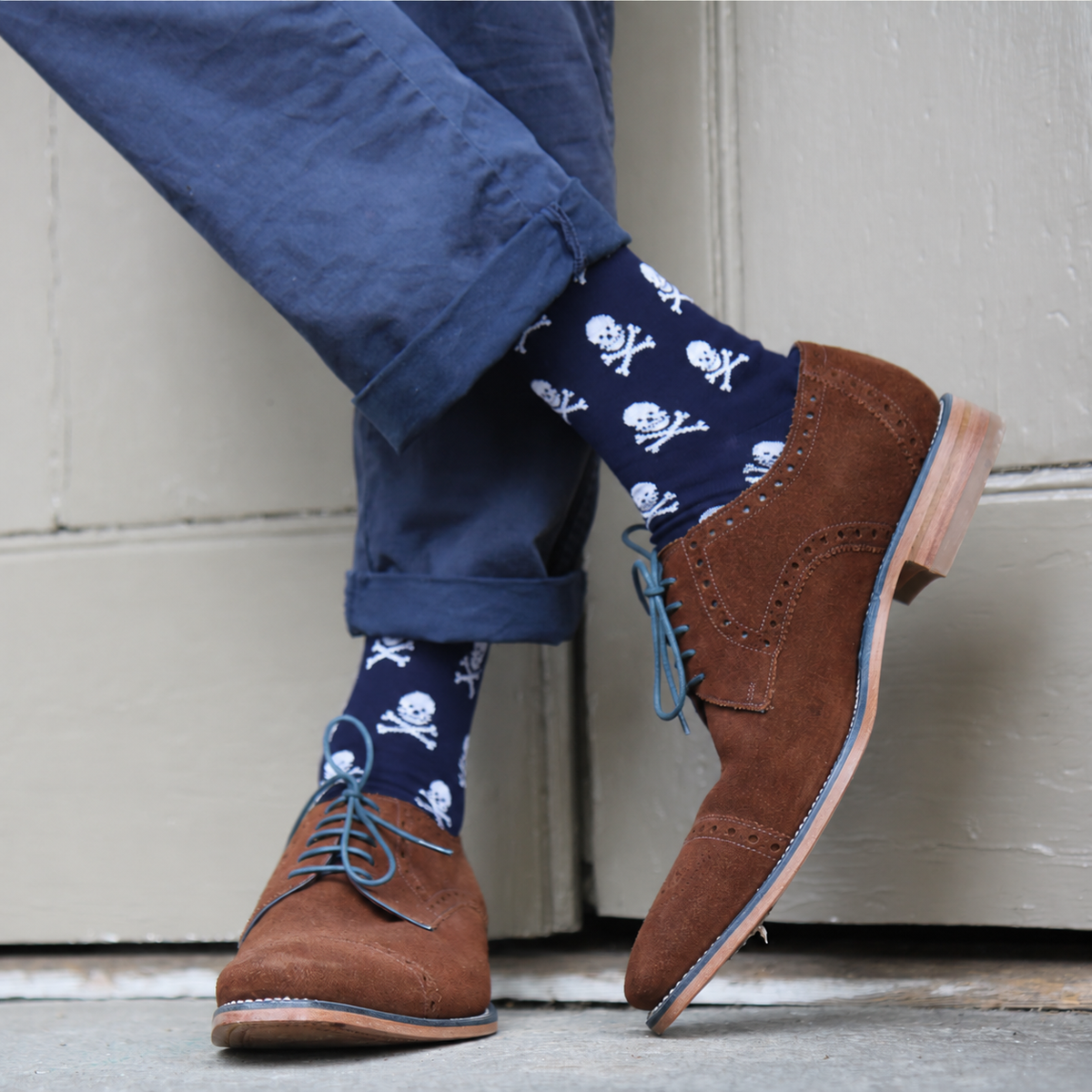 A person wearing navy blue socks with a white skull and crossbones pattern, paired with brown suede oxford shoes and navy pants, standing against a light gray door.