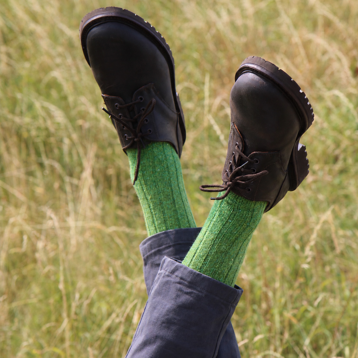 A person's legs kicked up in a grassy field, wearing bright green ribbed crew socks, dark brown lace-up boots, and dark blue jeans.