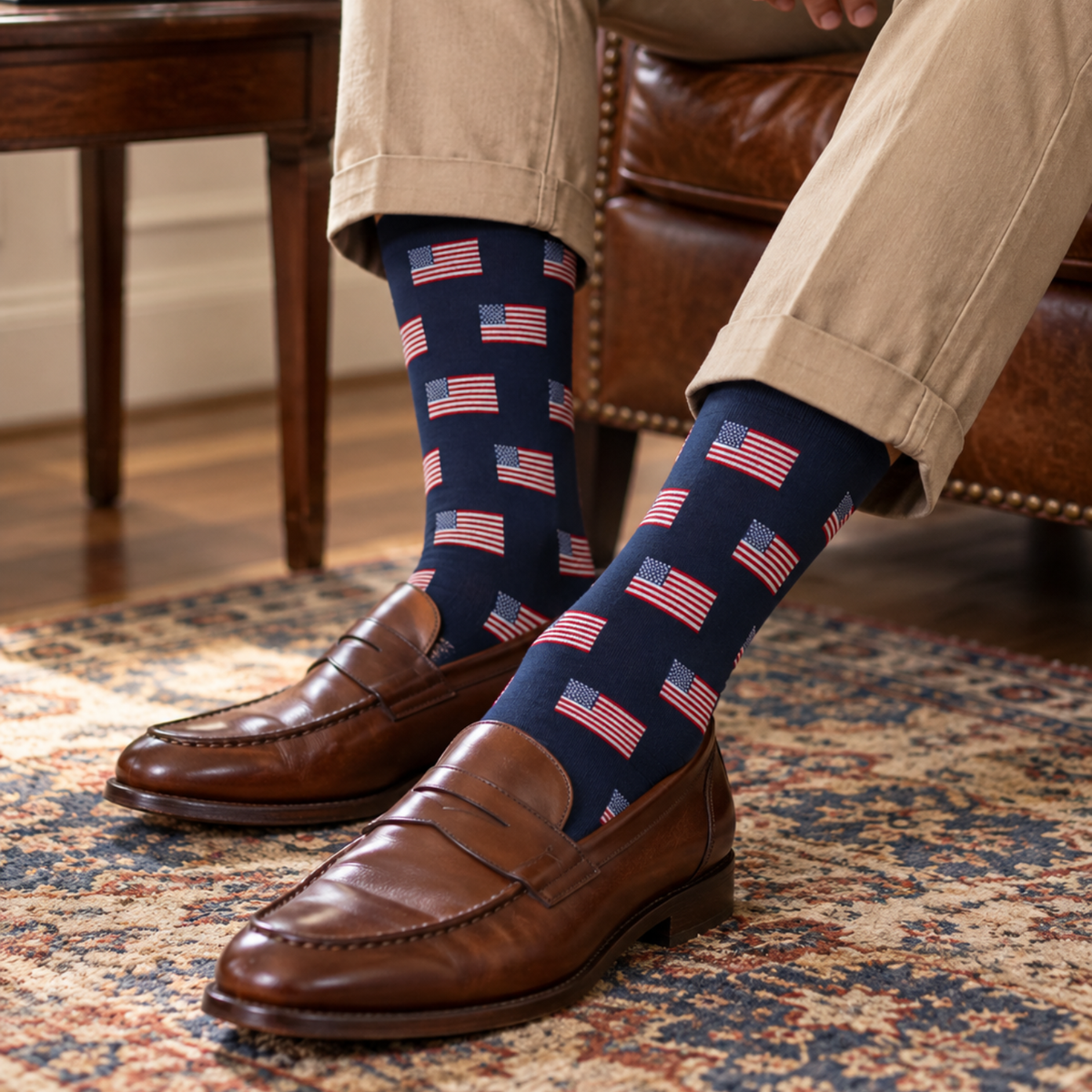 A person seated in a brown leather armchair wearing navy crew socks with a repeating American flag pattern, paired with tan trousers and brown leather penny loafers on a patterned rug.