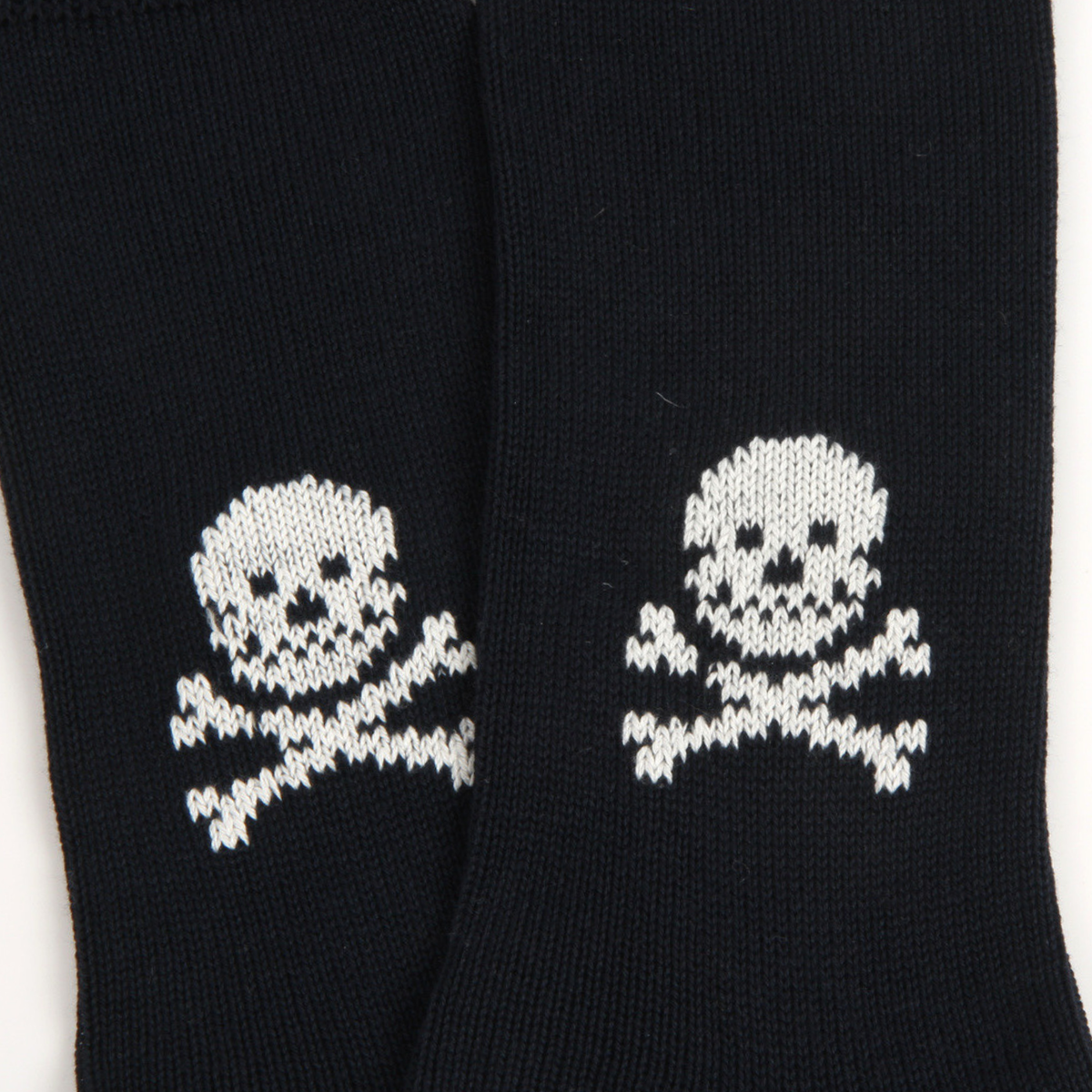 A close-up of a pair of navy knit socks featuring a white knitted skull and crossbones pattern on the lower leg.
