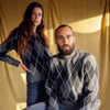 Two people wearing argyle sweaters sitting against a beige curtain.