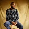 Man wearing a gray argyle sweater sitting on a wooden stool against a beige backdrop