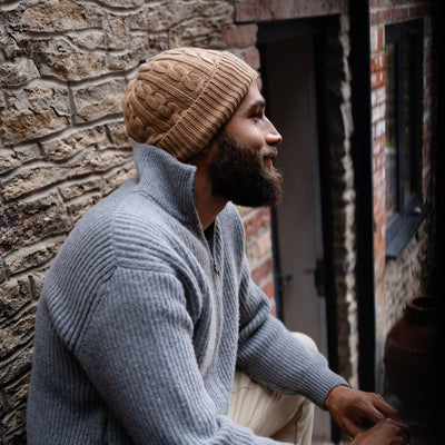 man wearing a cable cashmere beanie in colour brown sugar sitting outside on a step.