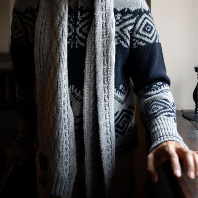 A man wearing a cable knit scarf with geometric patterns in navy, grey, and blue colours; the scarf is draped over his shoulders and the rest of the shirt and pants are not visible.