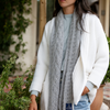 A woman wearing a white knit sweater and blue jeans, standing outdoors with a grey cable-knit cashmere scarf wrapped around her neck.