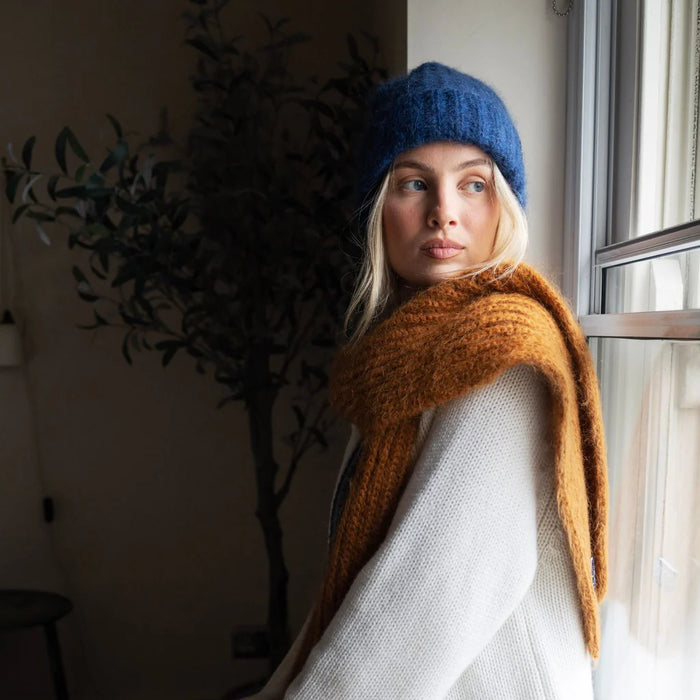 Woman wearing a blue knit hat and gold scarf indoors by a window