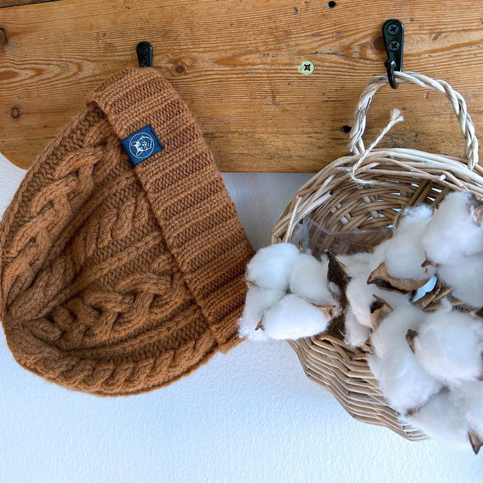 Brown knitted beanie with a logo tag next to a wicker basket filled with cotton on a wooden surface.
