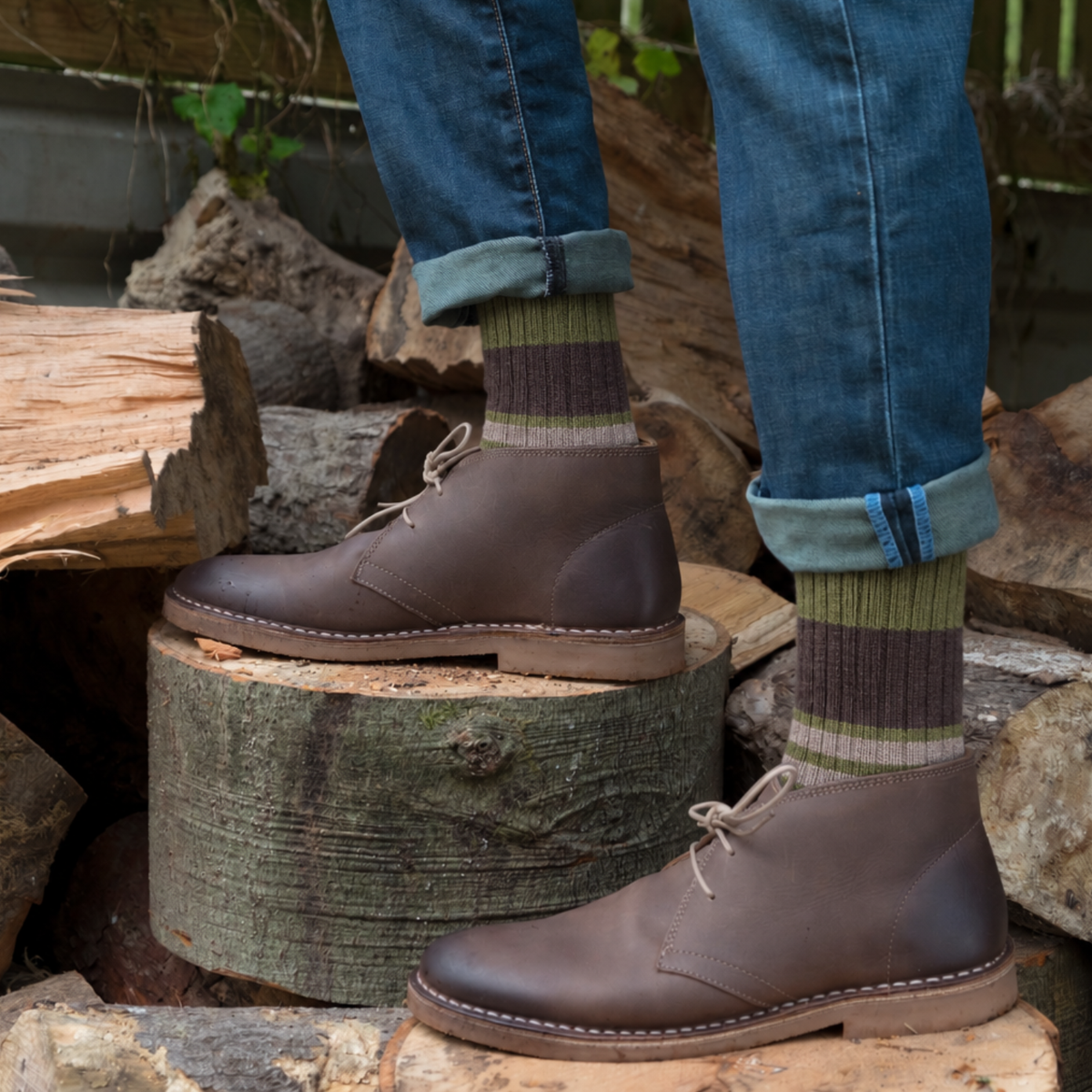A person wearing dark brown leather chukka boots, blue jeans with cuffed hems, and striped socks stands on stacked wood logs outdoors.