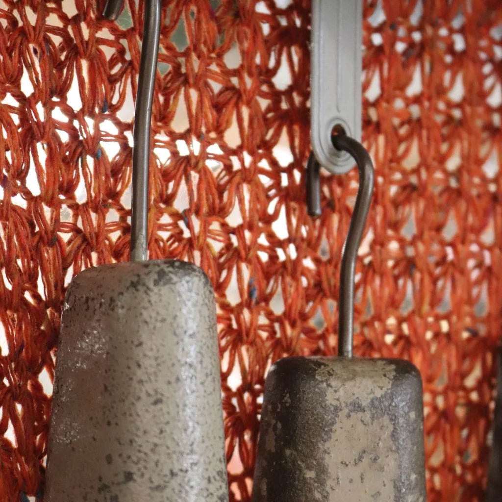 Close-up of rustic metal hanging weights suspended in front of a bright orange woven mesh curtain.