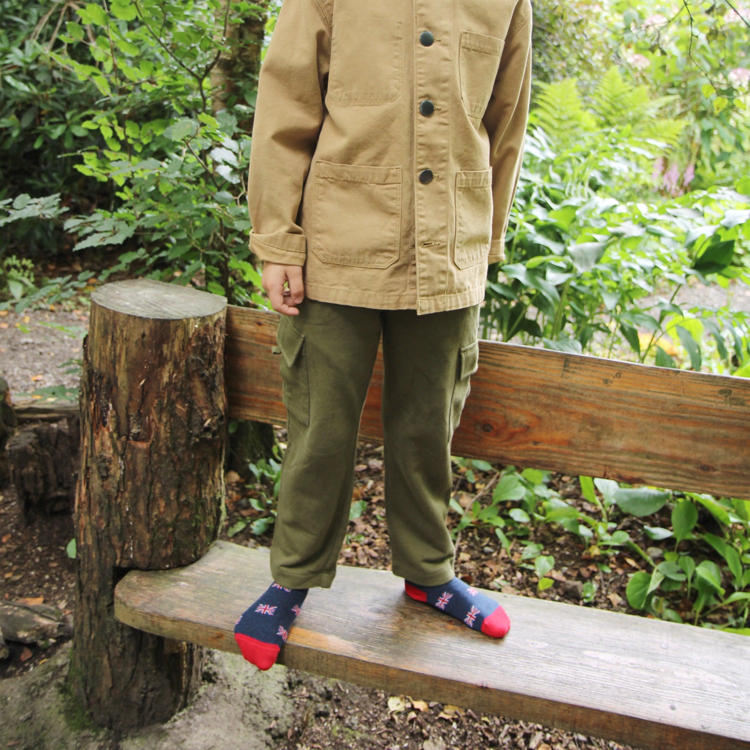 Child wearing a beige jacket and green pants sitting on a wooden bench in a forest setting.